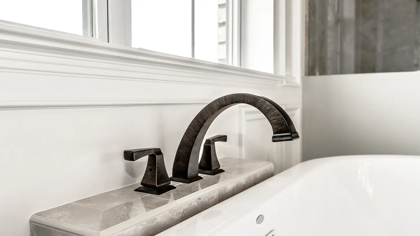 Close-up of a luxury bathroom tub deck featuring a brushed bronze curved faucet with matching handles, set against a marble surround and white wainscoting, designed by Frazier Homes.