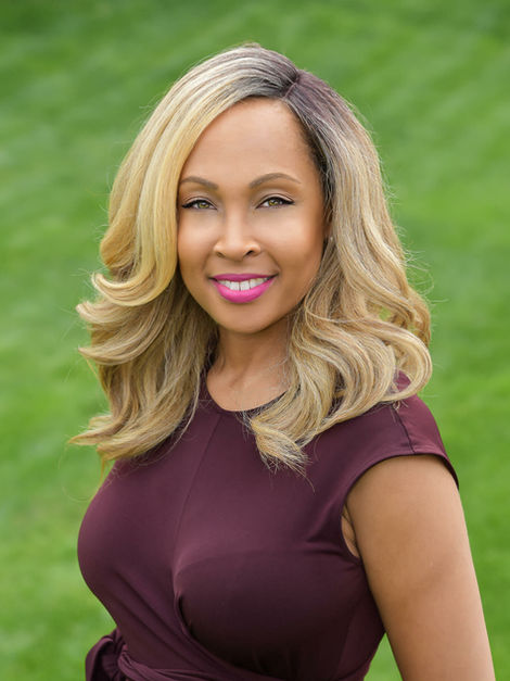 Professional portrait of Adrienne Frazier of Frazier Homes, wearing a burgundy dress and smiling outdoors against a green background.