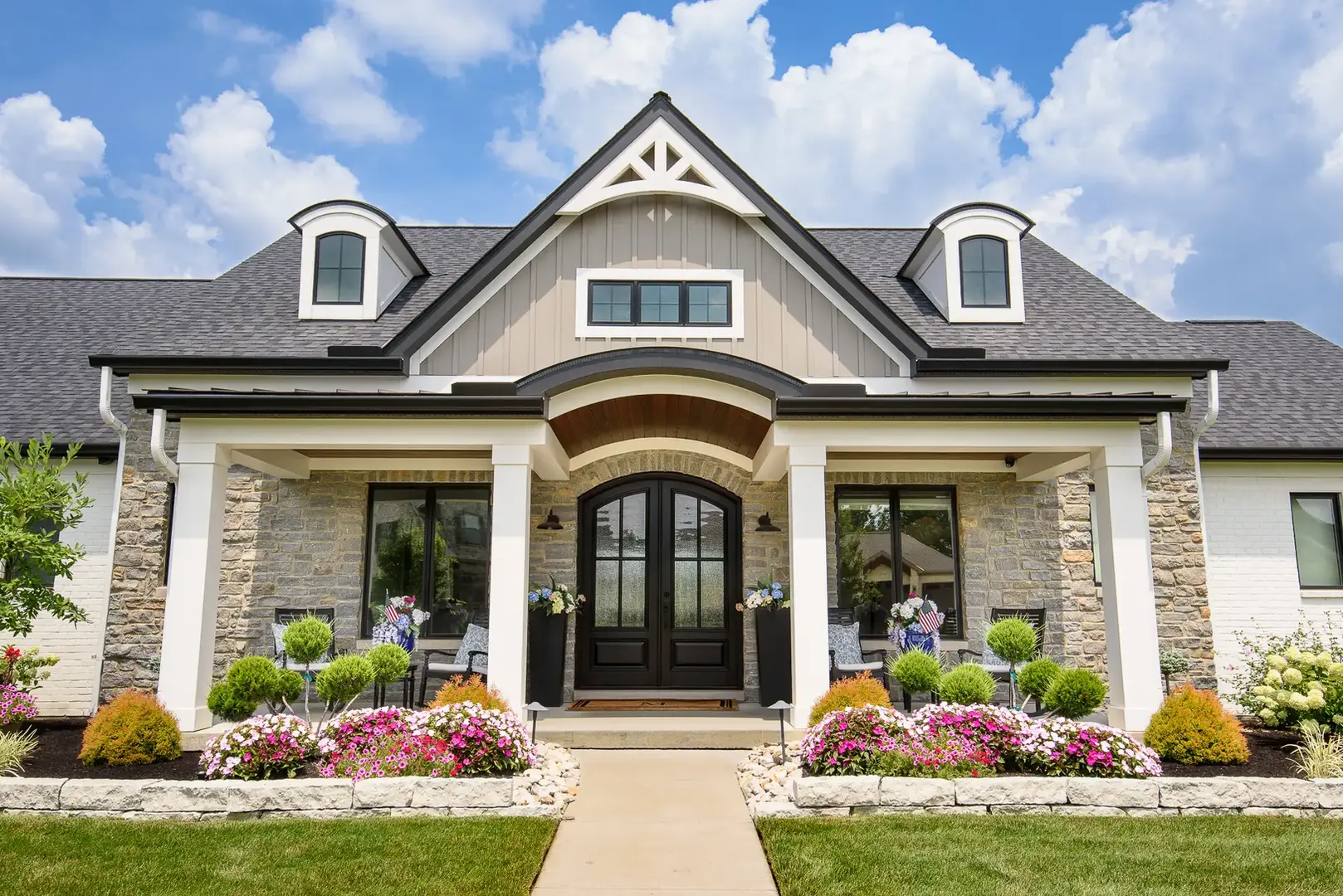 Luxury modern farmhouse exterior in Mason, Ohio by Frazier Homes, featuring stone and board-and-batten siding, an arched wood-lined entryway, black double front doors, dormer windows, white columns, and beautifully manicured landscaping.