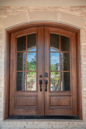 Close-up of arched double wood front doors with textured glass panes on a brick home exterior.
