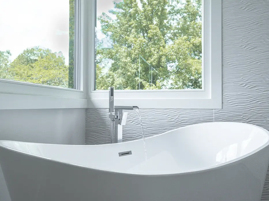 Freestanding white soaking tub with modern floor-mounted faucet in a Montgomery, Ohio luxury custom bathroom by Frazier Homes, featuring textured wall tile and large windows.