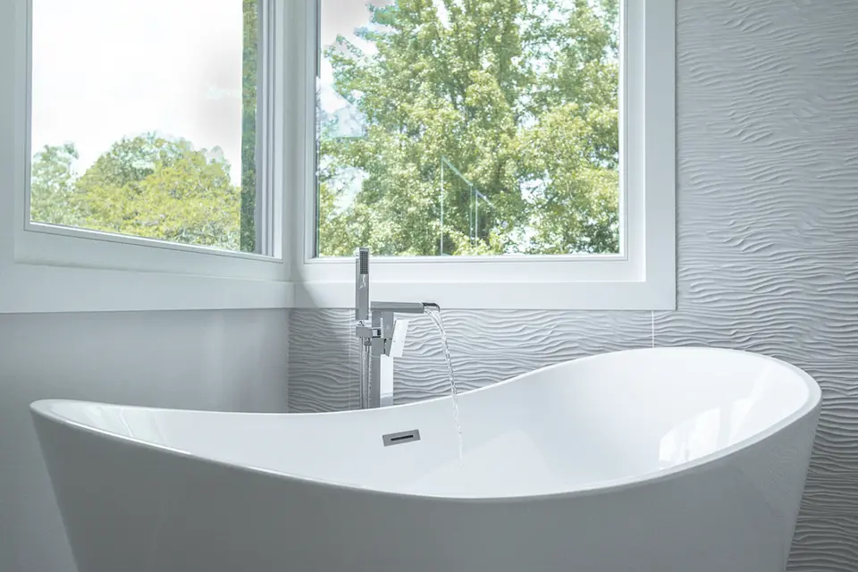 Freestanding white soaking tub with modern floor-mounted faucet in a Montgomery, Ohio luxury custom bathroom by Frazier Homes, featuring textured wall tile and large windows.