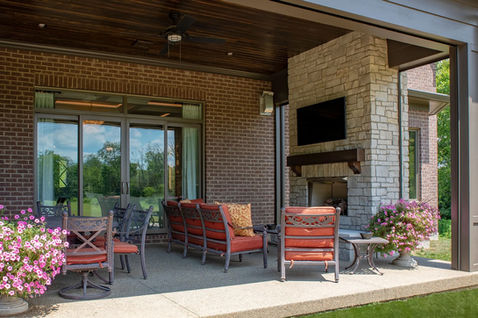 Covered patio with red cushioned seating, stone fireplace with TV, and blooming flower planters.