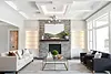 Luxury living room in Liberty Township custom home featuring a floor-to-ceiling stone fireplace with wood mantel, coffered ceiling with recessed lighting, built-in illuminated shelving, large picture windows, and modern white and black sofas arranged around a glass coffee table.