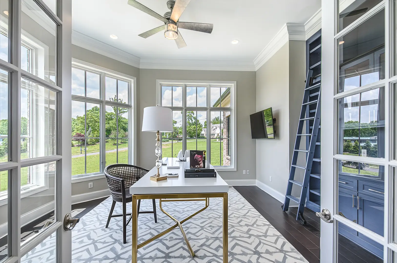 Bright luxury home office with floor-to-ceiling windows, white and gold desk, custom navy built-ins, and rolling library ladder in a Frazier Homes custom build.