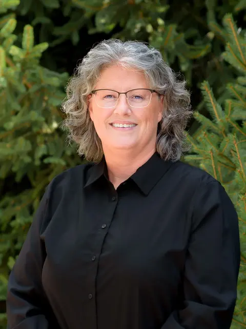 Professional headshot of an older woman with curly gray hair wearing glasses and a black blouse, standing outdoors in front of green trees.