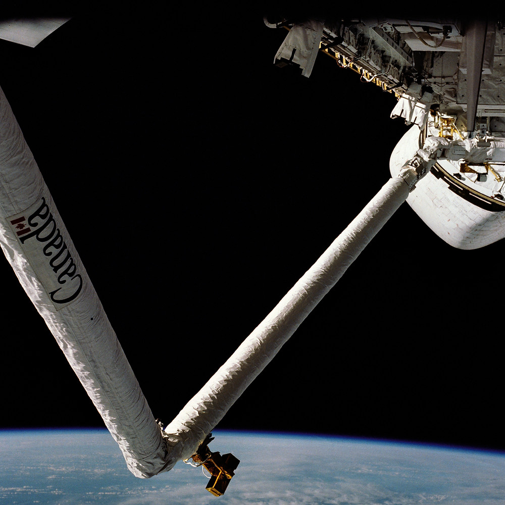A very long robotic arm extends from the space station. The Earth can be seen below the arm.