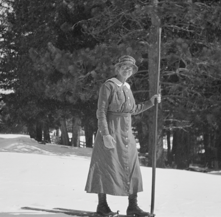 Photograph of Ynés holding a very tall walking stick and wearing cross country skis on a hillside.
