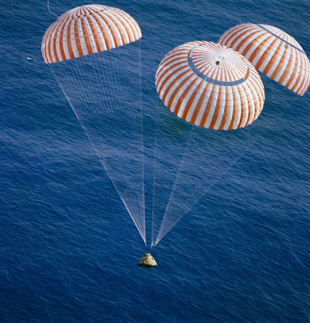 Photo of three parachutes attached to a small, cone-shaped vehicle suspended over an ocean.