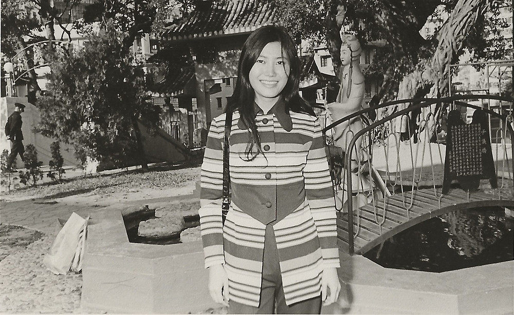 Flossie wears a striped coat and stands in front of an decorative garden with a fountain and bridge in Hong Kong.