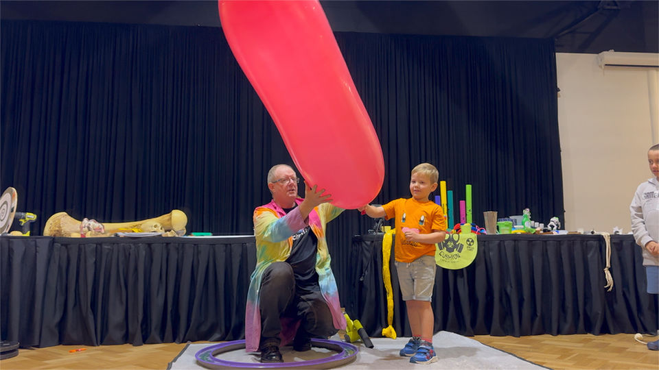 Child and science presenter performing giant balloon air pressure experiment on stage.