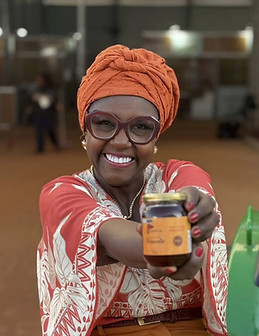 Mara, fundadora da Danega Geleias, sorridente, usando turbante laranja e roupa estampada, segura um pote de geleia da marca.