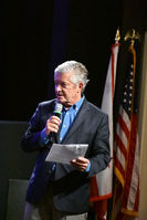 Gray-haired man speaking into microphone, American flag behind him