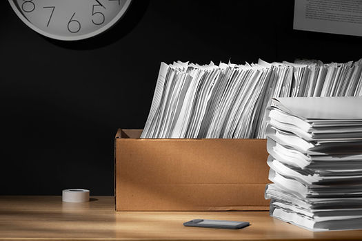 Overloaded desk with a box of papers and a tall stack of documents