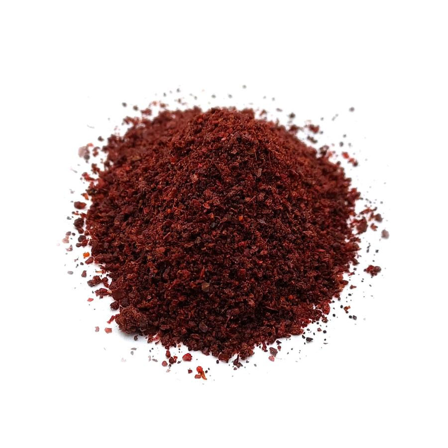 Сумах подрібнений, темно-червоний порошок спеції без упаковки на білому фоні.