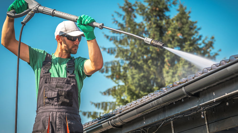 Man in green shirt and gloves pressure washes roof tiles under clear blue sky, surrounded by trees, concentrating intently.
