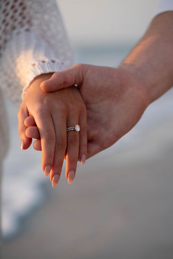 Close-up of couple holding hands on Florida beach with engagement ring in focus and cinematic ocean backdrop.
