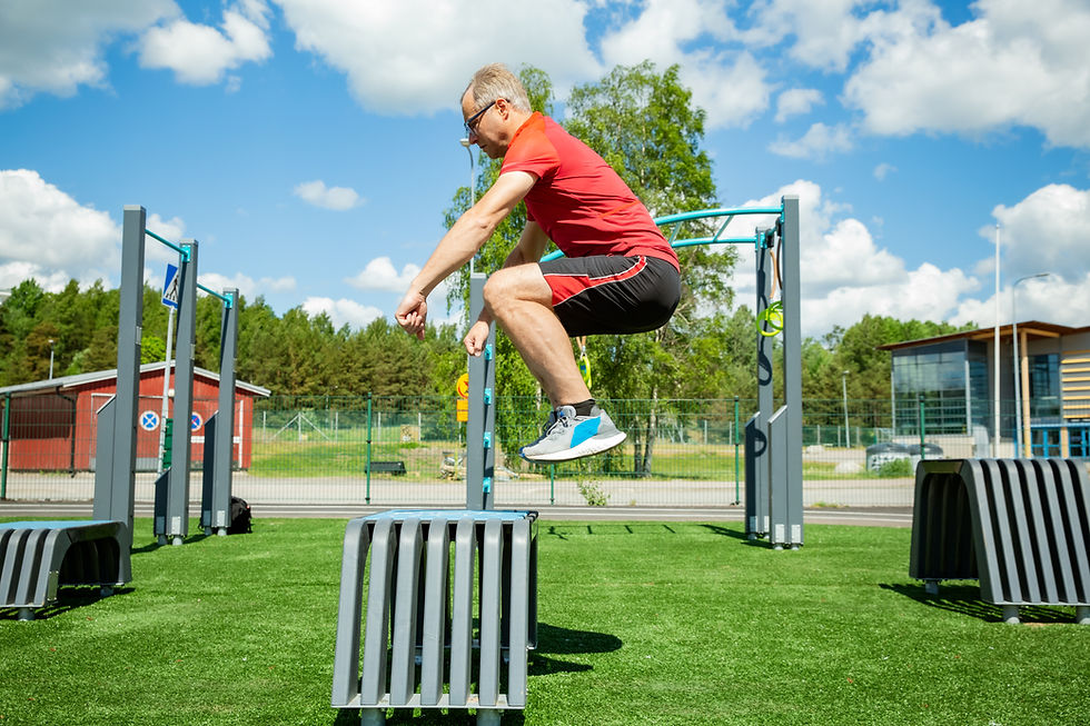 Man in red shirt and black shorts jumps over metal structure outdoors. Green field, blue sky with clouds. Athletic and energetic mood.