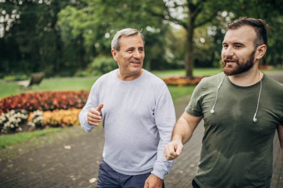 Two men walking and talking in a park. One wears a gray sweater, the other a green shirt with earphones. Colorful flowers in the background.