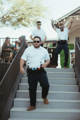 Groomsmen stride down the deck stairs with full beer holster belts at Windmill Winery, sunglasses and string lights signaling the loudest grand entrance.
