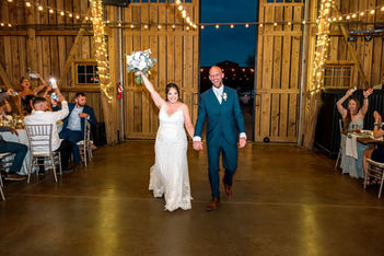 The newlyweds burst into Windmill Winery's barn through string-lit double doors, bouquet thrust skyward, guests cheering from the chiavari-lined tables on either side.
