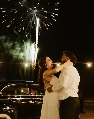 Bride and groom laughing and holding each other beside a vintage car at Windmill Winery while fireworks burst in the night sky behind them during their wedding send off.