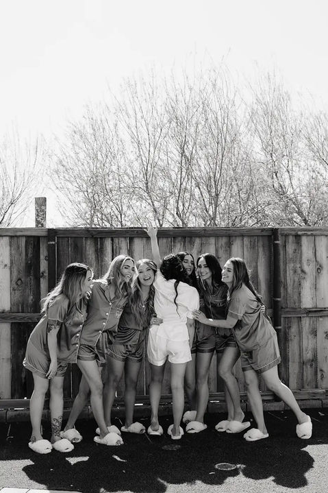 A bride and her bridesmaids gather at the exterior wall of Windmill Winery's barn beneath bare branches, in a black-and-white group portrait.