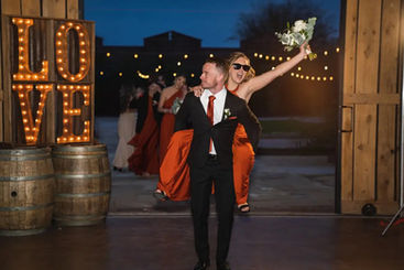 Newlyweds make their grand entrance through Windmill Winery's barn doors at dusk, bride leaning on groom's back beside the glowing LOVE marquee and barrels.