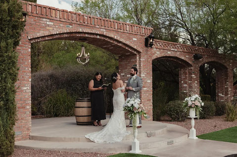Bride and groom exchange vows at Windmill Winery's brick aqueduct ceremony arch, white floral arrangements and wine barrel setting the scene at golden hour.