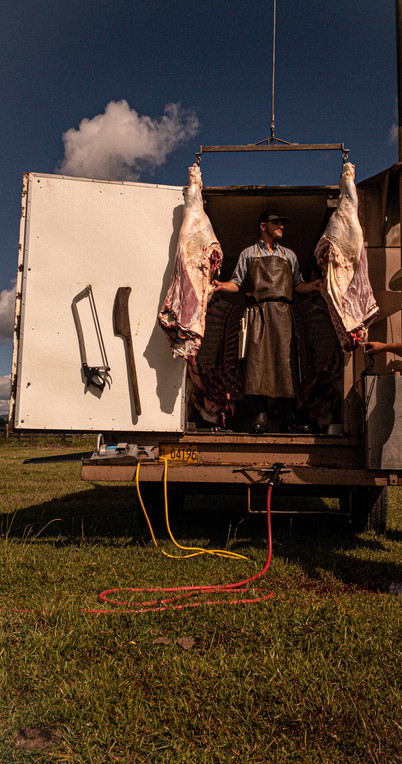 A white man in his mid-20s is wearing a black butcher's apron. He is standing in between two pork carcasses. To the left of the image are two butchering instruments: an ax and a saw. The sky is dark blue.