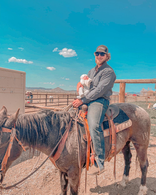 A photo of a white man with hair to his ears wearing black sunglasses and a ball cap. He is sitting in a saddle on a light brown horse. In his left hand, he holds the horse's reigns. In his right hand, he holds a newborn baby who is wearing a white hat and a white outfit. There is brown fencing behind them.