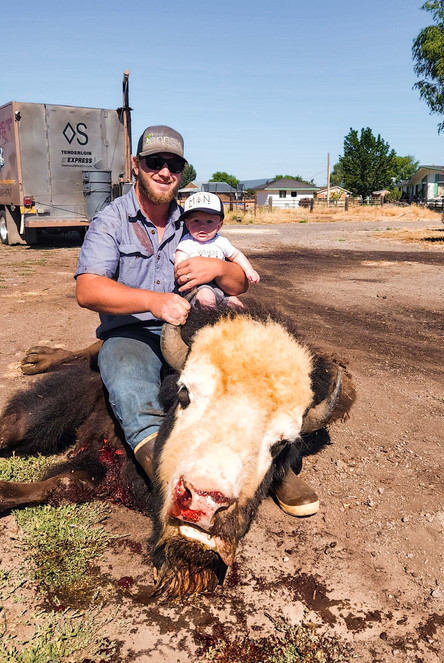 A white man with hair to his ears wearing black sunglasses and a ball cap is holding a 4 or 5 month old baby who is wearing a white shirt and a white ball cap. They are both sitting on the back of a butchered "beefalo - half buffalo, half cow. The beefalo's head is white and it has black eyes and brown horns. There is blood coming out of its mouth and nose. A silver metal butchering trailer stands in the background. 