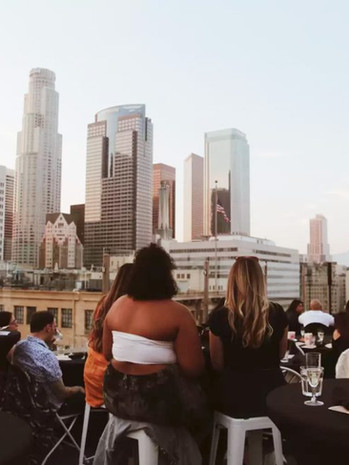 Outdoor rooftop event with people seated at tables overlooking the downtown city skyline at sunset.
