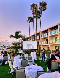 Outdoor event setup with round tables and chairs on grass, palm trees, and a hotel building at sunset.