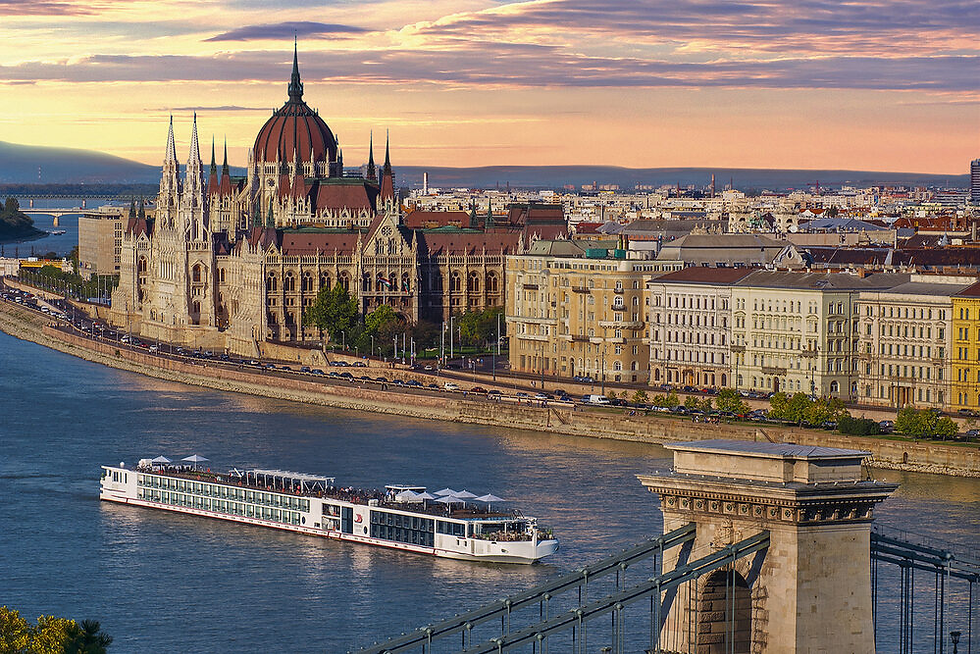 White river cruise ship cruising along the Danube River in Budapest, with the Hungarian Parliament Building and historic city architecture lining the waterfront at sunset.
