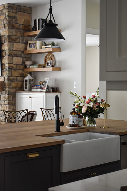 Oak countertop on island with dark flush inset shaker cabinetry, black faucet and fresh flowers, overlooking stone fireplace in livingroom with custom builtins, floating shelves, white cabinetry made in Ontario with Ontario hardwood