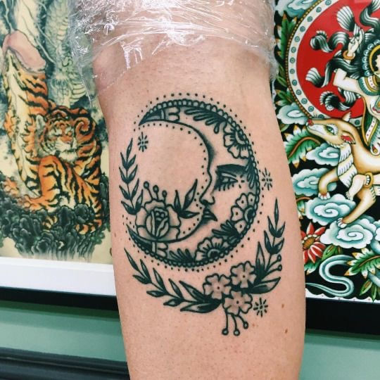 traditional black and grey linework moon tattoo with small flowers on womans lower leg