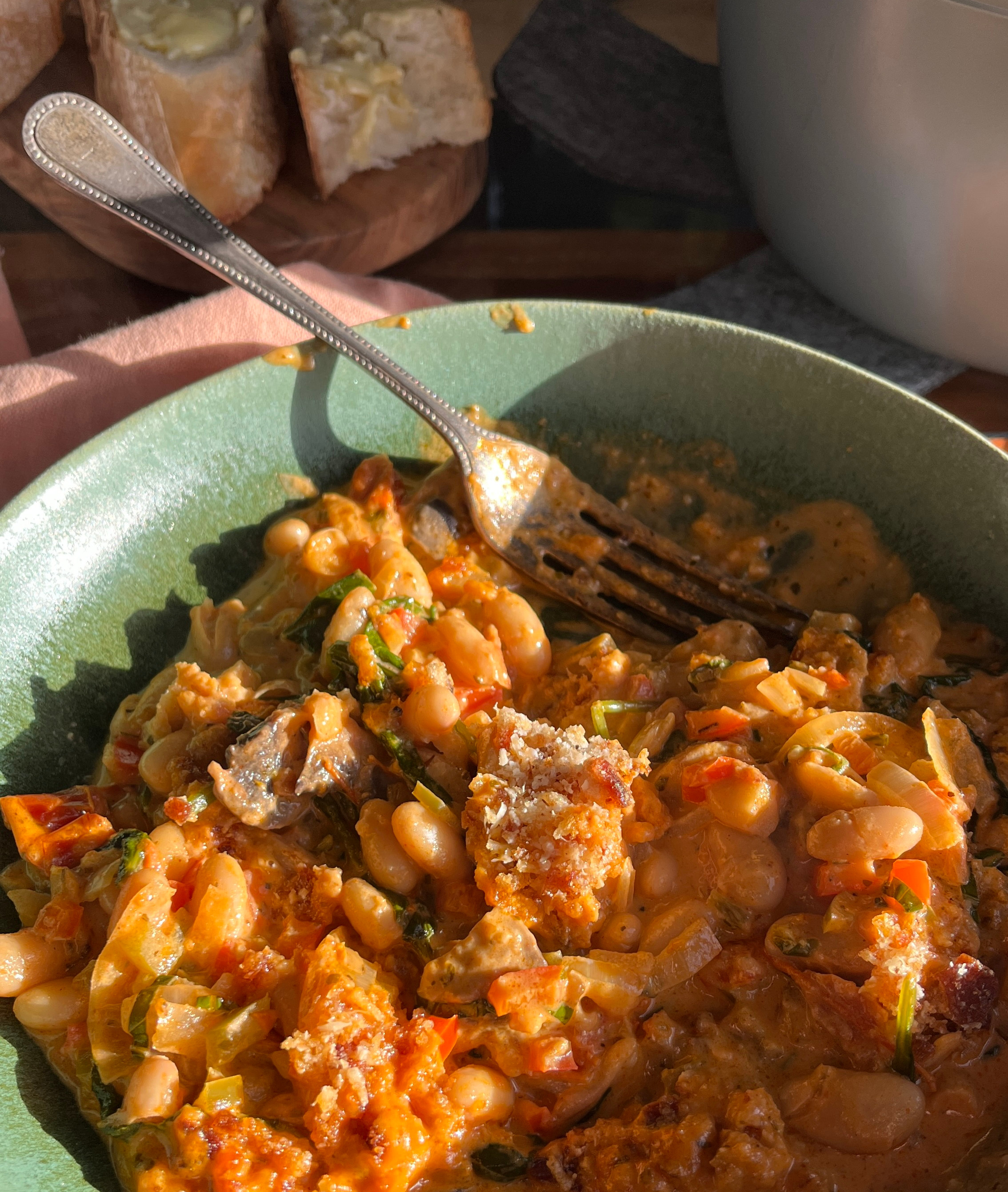 A large bowl of creamy tuscan bean stew sits on a table top; it is warm and orange in colour. There is a crisp breadcrumb topping.