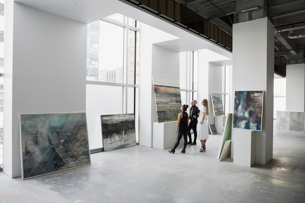 Three people stand in spacious warehouse looking at oil paintings