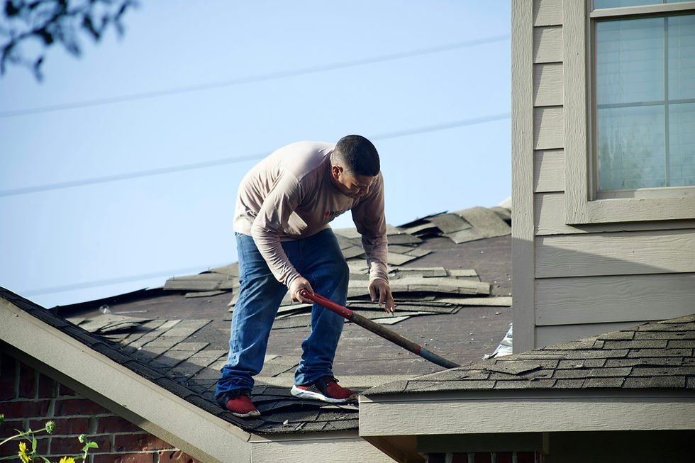 Man repairs roof with a tool, surrounded by shingles. Wears jeans and red shoes. House exterior visible with blue sky.