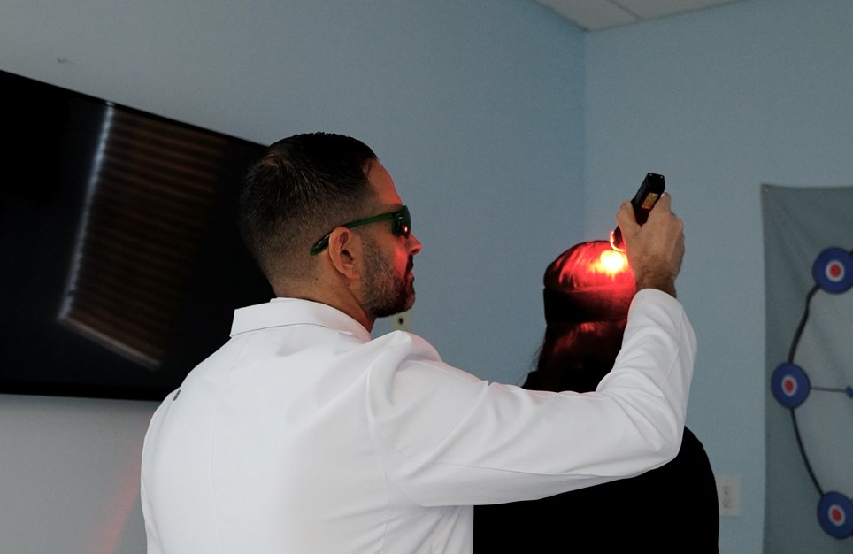 A person in a white coat uses a laser device on another's head in a blue room with diagrams. The mood is clinical and focused.