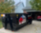 Black dumpster labeled "RHINO Roll Off Dumpster Rental" on driveway by garage with red car. Leaves on ground; tree and house visible.