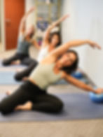 Three women in workout gear stretch with exercise balls in a calm room. They're on yoga mats, with a shelf of folded blankets behind them.