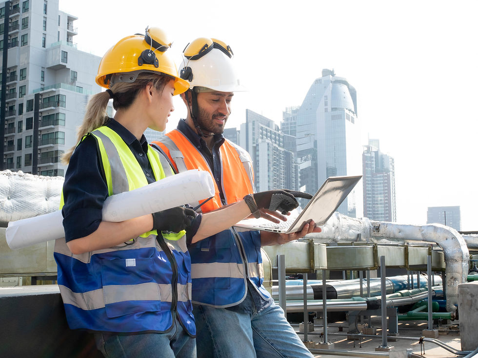 Two construction engineers in hard hats and safety vests review a laptop and blueprints on a rooftop with city skyscrapers behind.