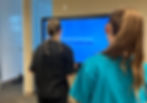 Two people in a room stand before a blue screen displaying "Find the following words in reverse sequence." One wears black, the other teal.