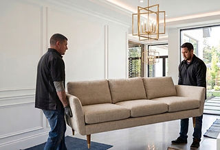 Two men move a beige sofa in a modern living room with white walls and gold pendant lights. Large windows reveal a garden outside.