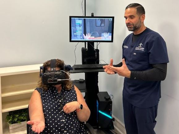 A woman in a polka dot shirt wears a VR headset, sitting next to Dr Conde of Functional Neurology Brain Center of Florida in blue scrubs who gestures. A screen shows virtual hands.