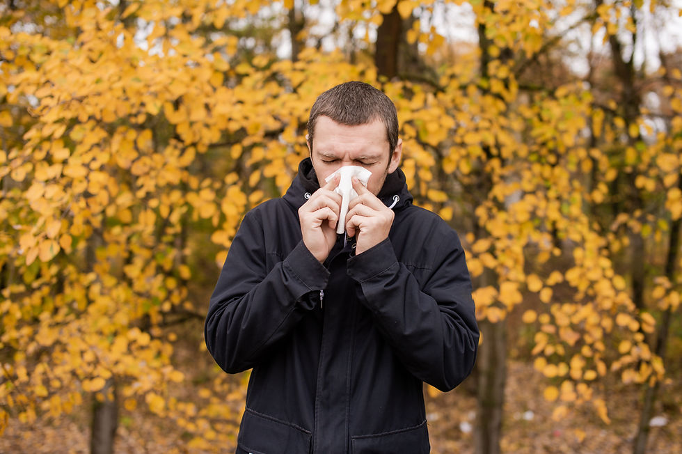 Man in black jacket sneezing into tissue, stands in front of yellow autumn leaves. Looks chilly and unwell, outdoor setting.