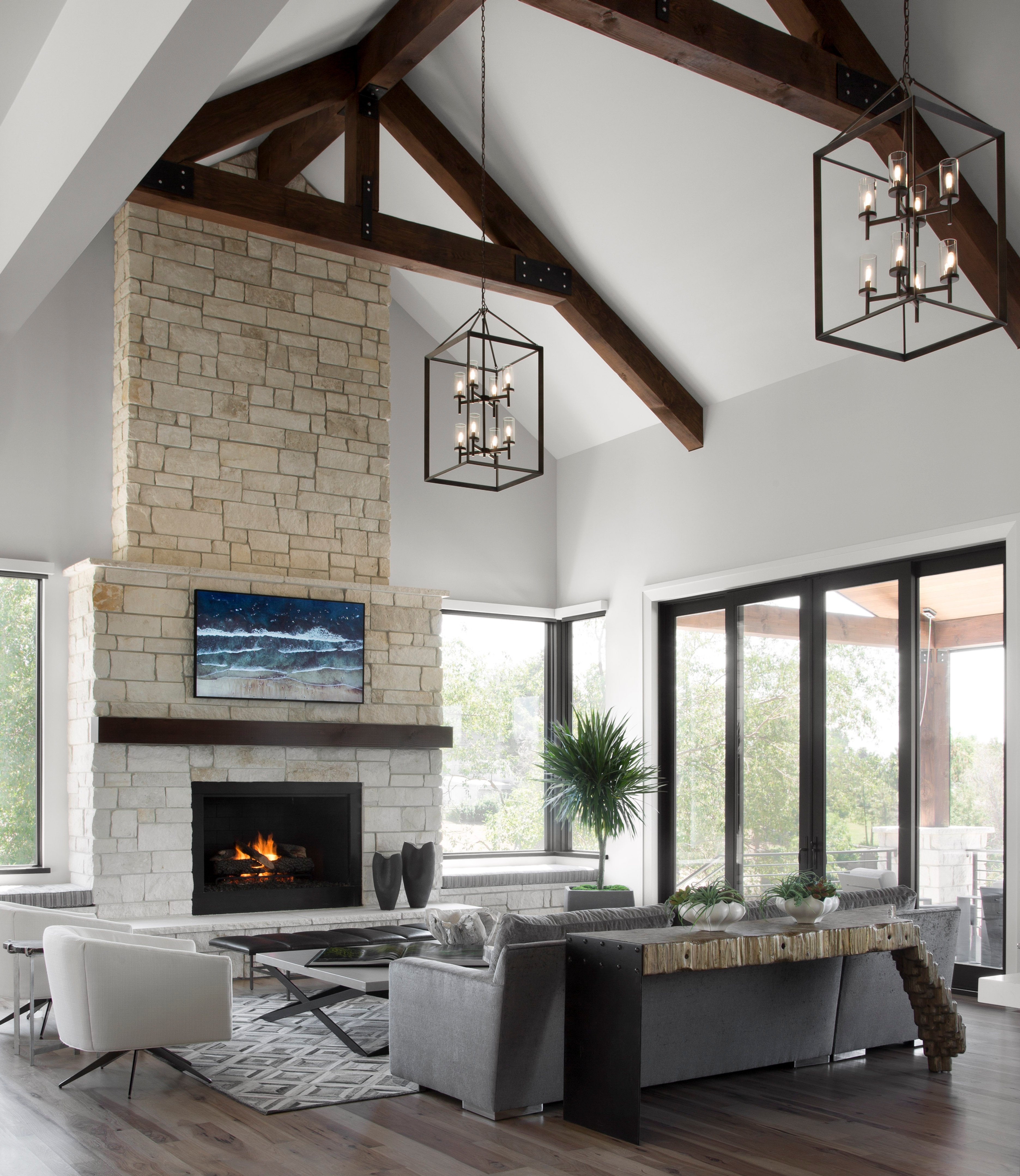 Bright modern living room with stone fireplace, lit TV above, gray sofas, wood beams, hanging lanterns, and large windows.