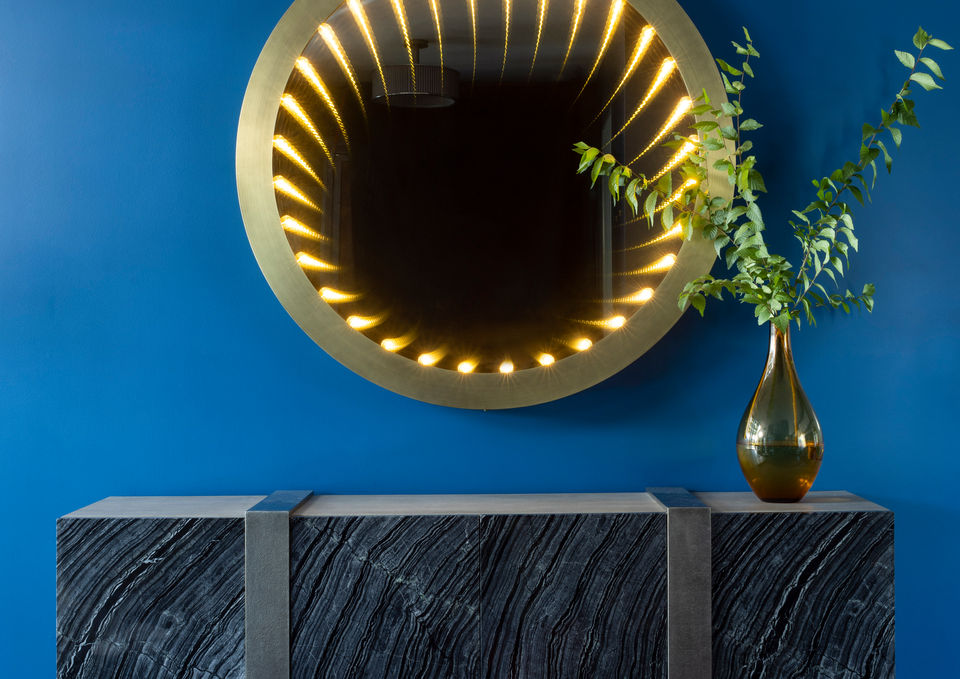 Round mirror with lights above a gray cabinet against a blue wall.